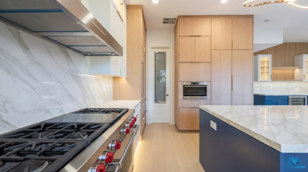 Pro range with red knobs below a marble-look backsplash, with light wood tall cabinets and a blue island, San Jose